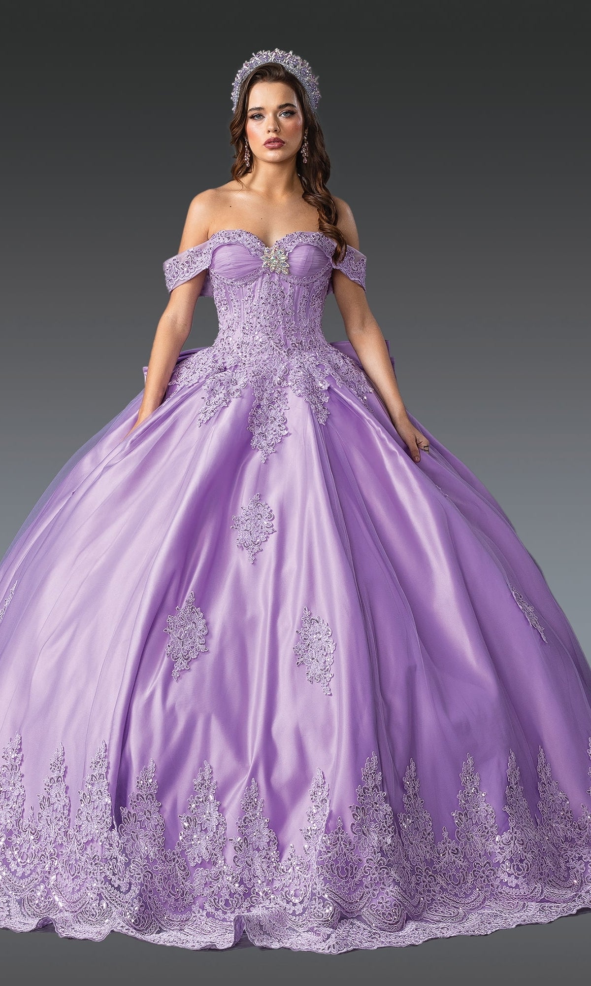 A woman in the Quinceanera Dress 1913 By Dancing Queen, featuring an off-the-shoulder lavender design with a corset bodice, lace details, and a matching tiara, stands against a gray background.